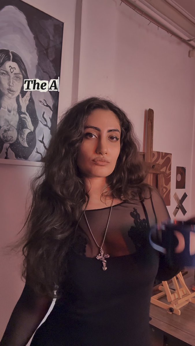 A woman with long dark curly hair wears a cross necklace and stands in a room with artwork and a wooden easel, with text 'The A' visible.