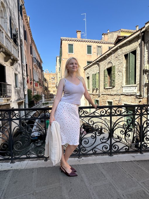 A blonde woman in a white sleeveless top and a white polka-dot skirt stands on a bridge in what appears to be Venice, holding a jacket, with canal-side buildings behind her and a 'FONDAMENTA S. POLO' sign visible.
