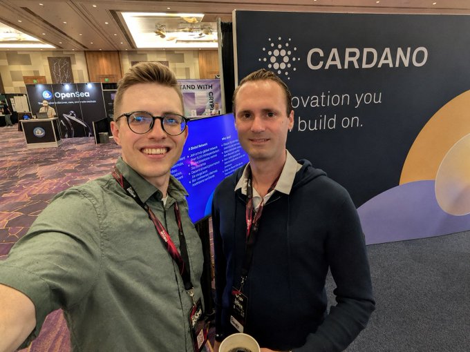 Two men smile for a selfie in front of a Cardano-branded booth that reads "Cardano – Innovation you build on," at the RareEvo 2026 conference. The accompanying post mentions David Kinitsky of Everstake speaking at the event and a warm handshake of community in the Web3 space.