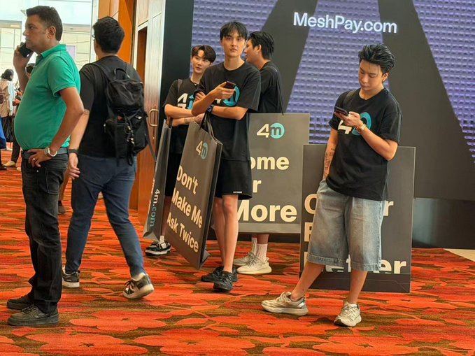 A group of young men in black "4D" branded t-shirts are standing in front of a MeshPay.com backdrop, with some holding promotional signs that read "Don't Make Me Ask Twice".