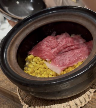 A black bowl contains a dish featuring raw red meat slices resting on a bed of yellow corn kernels.