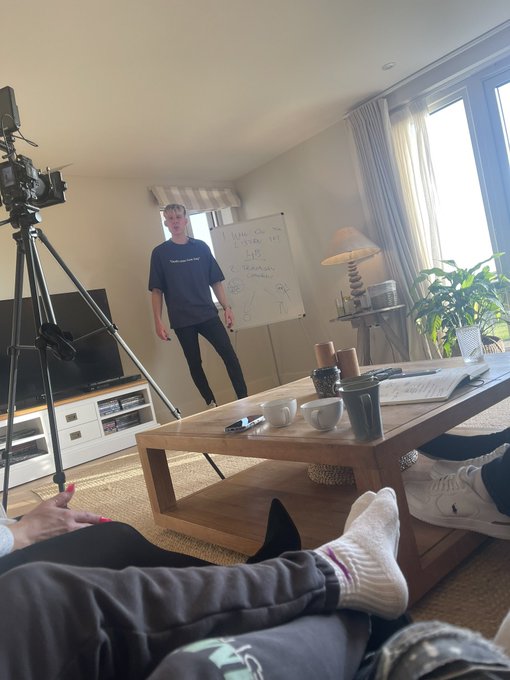 A young man in a blue t-shirt stands in a living room next to a whiteboard with handwritten notes including 'GROWTH ACCOUNT' and '2 TRADERS', while someone wearing socks sits on a couch in the foreground filming with a camera on a tripod.