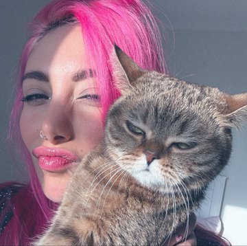 A close-up photo of a young woman with bright pink hair, a nose ring, and glossy lips posing beside a grumpy-looking tabby cat with green eyes.