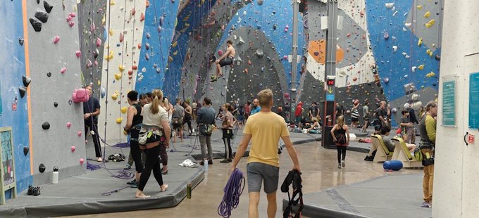 An indoor rock climbing gym with multiple colorful climbing walls and numerous climbers of various ages, including a man in a yellow shirt holding climbing rope in the foreground.