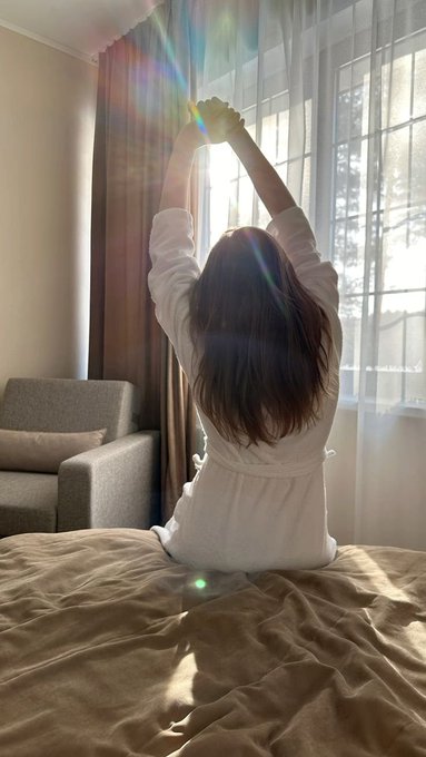 A person with long dark hair sits on a bed facing a bright window, stretching their arms upward with sun rays streaming through sheer curtains.