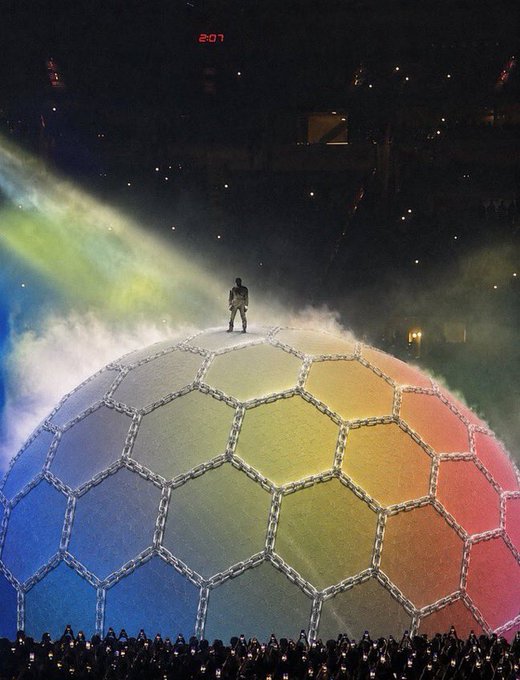 The user wants a caption describing the image, including any visible text.
The image shows a large, spherical structure resembling a soccer ball made of hexagonal panels.
The panels are colored in a rainbow gradient (blue, green, yellow, orange, red).
A person stands on top of the sphere.
There is smoke or mist around the base of the sphere.
In the background, there are stadium lights and a red digital clock showing "2:07".
The bottom of the image shows a crowd of people holding up lights (likely phones).
The prompt asks to relate this to the text: "The next wave of innovation on Solana won’t just be faster—it’ll be smarter. I’m excited to see how @world_xyz is empowering builders to launch projects that combine speed, scalability, and real utility. The winners won’t be the loudest projects. They’ll be the ones solving".

The image looks like a scene from a major event, possibly the Olympics (Be