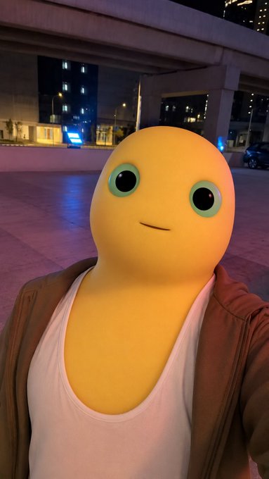 A yellow cartoon-like character with large green eyes and a slight smile, dressed in a white tank top and brown jacket, takes a nighttime selfie under an overpass with city lights in the background. The casual, quirky image plays into the lighthearted tone of seeking a fun, open-minded rating without being too serious.
