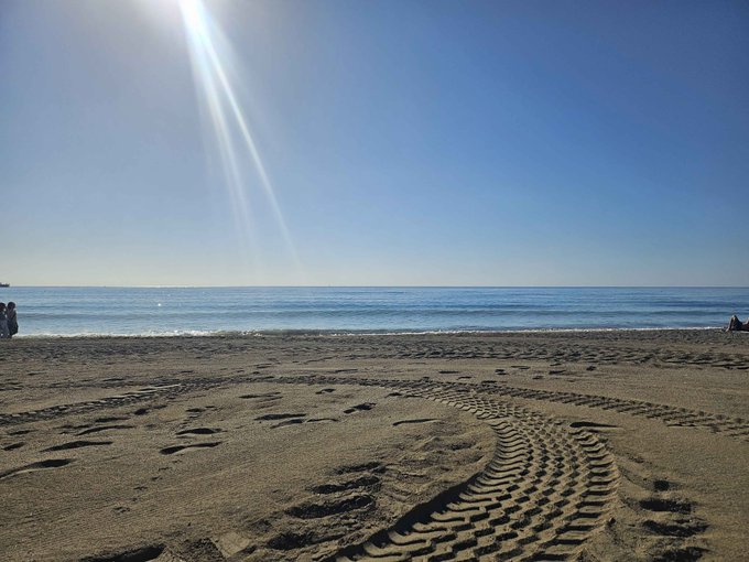 A sunny beach scene featuring sand with footprints and tire tracks in the foreground, leading to calm blue ocean waters under a clear sky with bright sun rays.