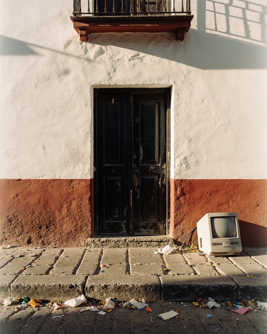 A weathered dark wooden door set in a white and red painted wall, with an old CRT television discarded on the cobblestone sidewalk beside scattered trash.