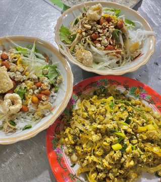 Three plates of Asian cuisine on a metal table, including rice topped with fried items and peanuts, a glass noodle salad with vegetables, and a yellow stir-fry.