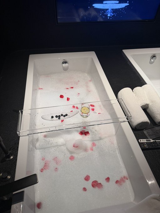 A white bathtub filled with foamy water and red rose petals features a floating tray holding a glass of yellow liquid and black beans, with a digital thermometer displaying "80" visible on the counter nearby.