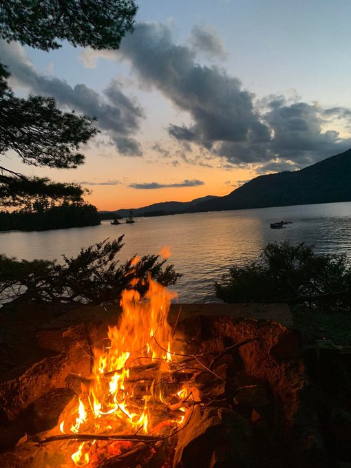 A scenic view of a campfire burning in a stone fire pit at sunset, overlooking a lake with mountains in the background and silhouetted trees framing the scene.