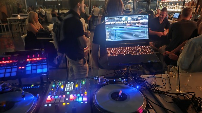 A DJ setup with illuminated equipment, a laptop showing mixing software, and people gathered around in what appears to be a bar or club setting.