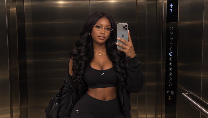 A woman taking a mirror selfie in an elevator, wearing a black Gymshark sports bra and matching leggings, with a '7' displayed on the elevator panel.