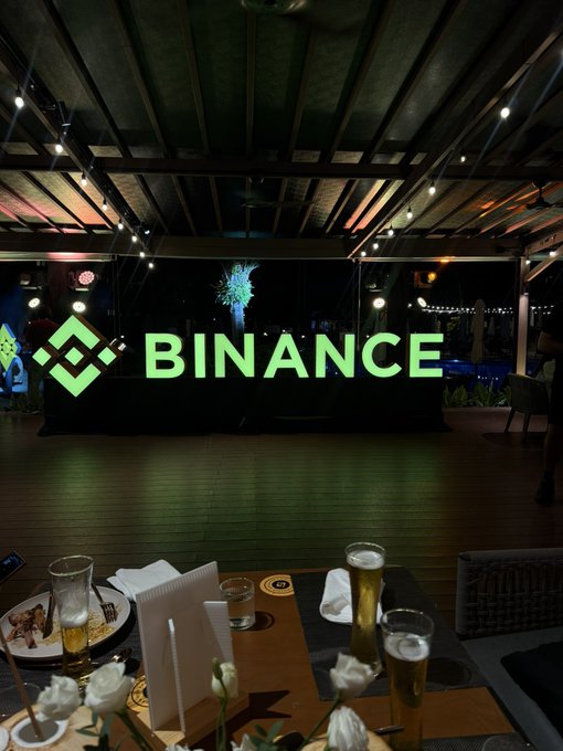 A welcome dinner event featuring the illuminated Binance logo on a stage, with a table set in the foreground holding beer glasses, floral arrangements, and dining ware. The scene captures the venue ambiance for the #BinanceCampusAsia gathering.