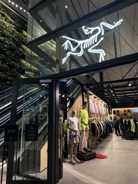 An Arc'teryx retail store interior with its iconic dinosaur skeleton logo illuminated on the glass wall, showing mannequins and apparel displays.
