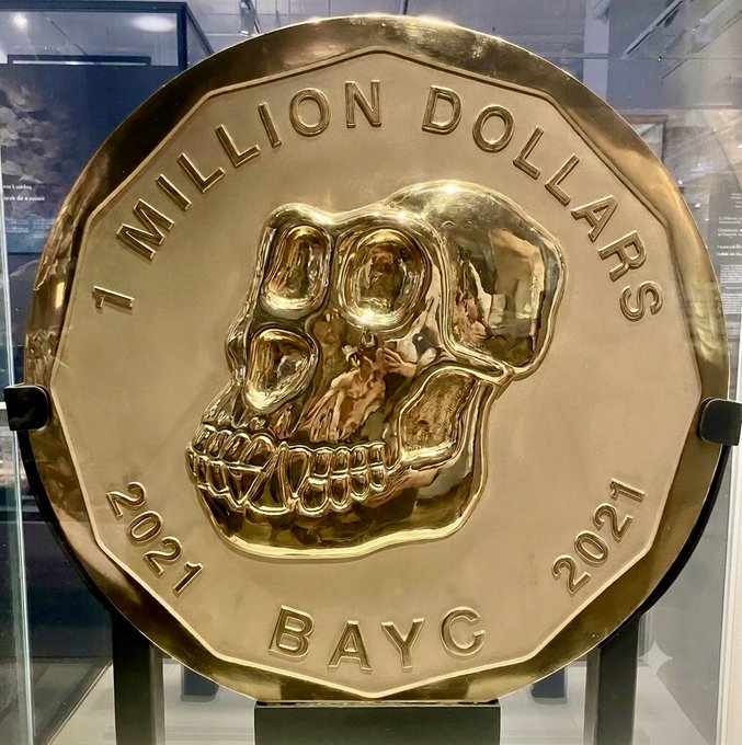 A large gold-colored commemorative coin displayed in a case, engraved with '1 MILLION DOLLARS', '2021', and 'BAYC' surrounding a skull design.