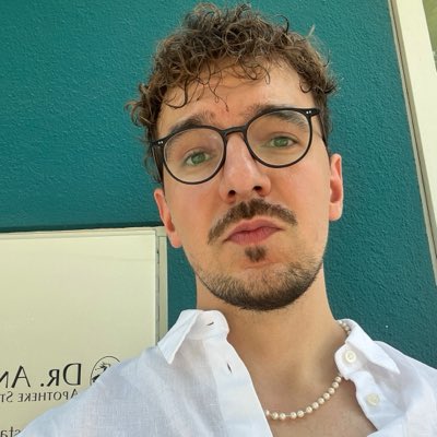 A close-up selfie of a young man with curly brown hair, glasses, a beard, and a mustache, wearing a white shirt and pearl necklace, standing in front of a teal wall.