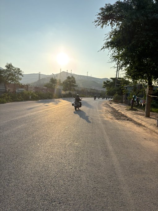 A motorcyclist rides down a sunlit rural road during golden hour, with the sun low in the sky and hills visible in the background.