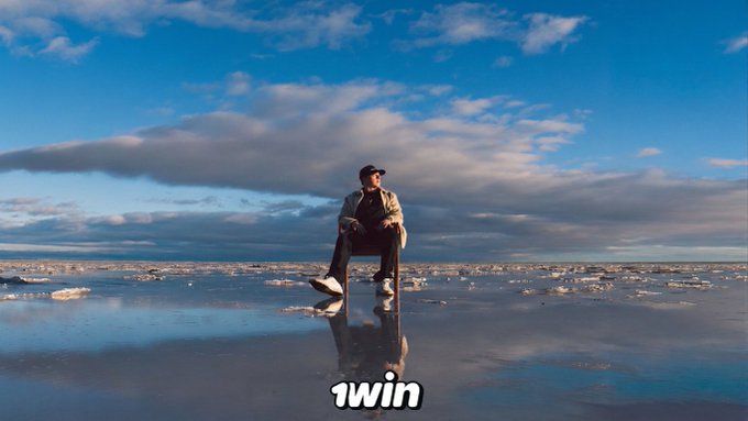 An edited movie scene showing a man sitting on a chair in shallow water on a beach under a blue sky, with the word '1win' displayed at the bottom.
