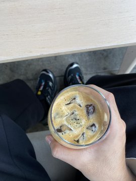 A hand holds an iced coffee in a clear plastic cup while sitting down, with sneakers visible below.