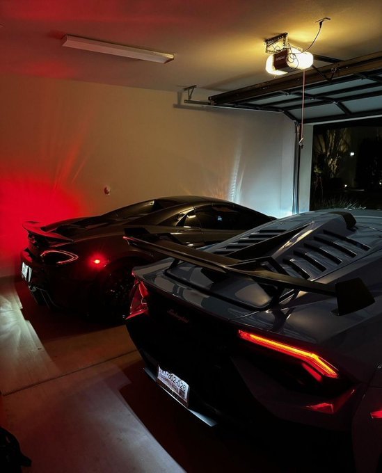 Two sleek black sports cars parked in a garage, one appearing to be a McLaren and the other a Lamborghini, illuminated by red ambient lighting and a ceiling lamp.