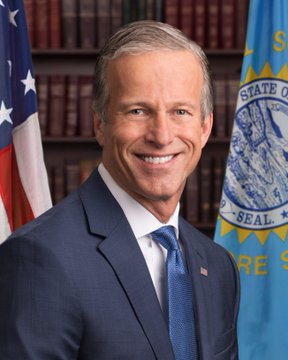 An official portrait of Senator John Thune, smiling in a blue suit, standing before the US flag and the South Dakota state seal.