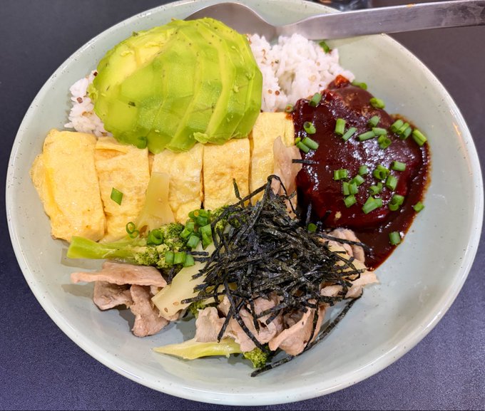 A bowl of rice topped with a fanned avocado half, tamagoyaki (rolled omelet), meat in dark red sauce garnished with green onions, shredded nori seaweed, and broccoli.