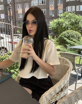 A young woman with long dark hair, wearing sunglasses and a cream-colored blouse, sips a drink while seated at an outdoor café. The post's cheerful message about Tezos Tuesday ties into the relaxed, sunny café setting, where the woman appears to be enjoying a lucky break with her beverage in hand.