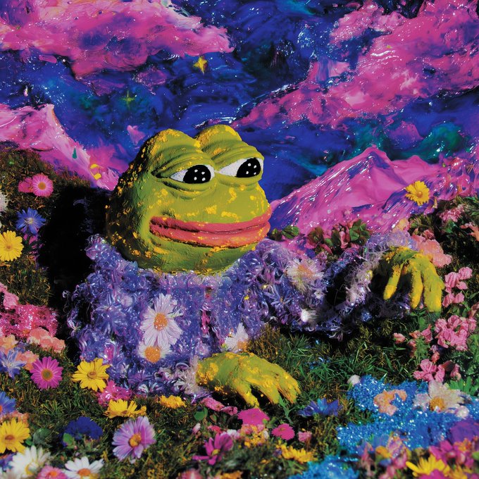 A textured, painted sculpture of a Pepe the Frog figure lying among colorful flowers, wearing a floral purple garment, set against a vibrant pink and blue sky.