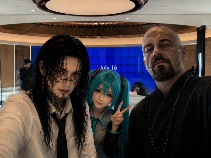 A selfie of three people in a modern room: a person with long black hair and facial tattoos, a cosplayer dressed as Hatsune Miku with teal twintails, and a bald man wearing a black robe and beaded necklace, with 'July 16' displayed on a screen behind them.