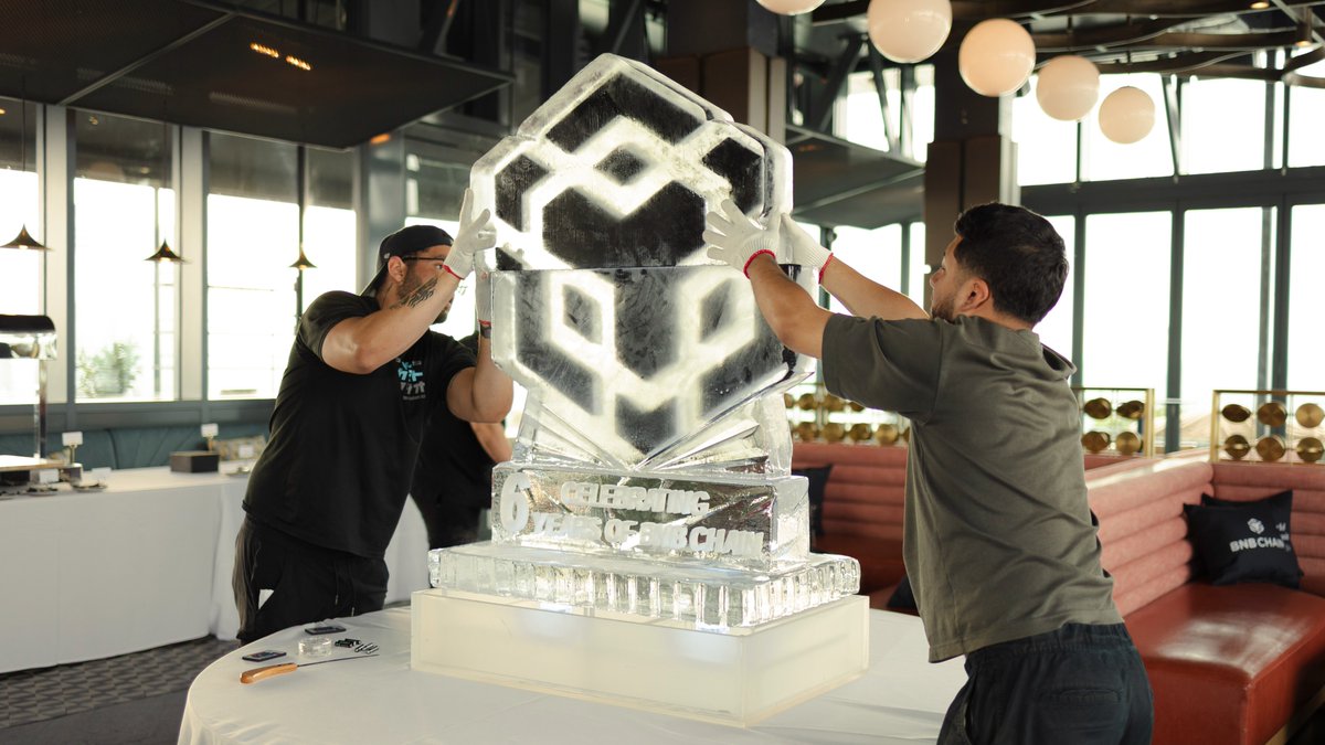 Two workers carefully assemble an intricate ice sculpture featuring the BNB Chain logo and the text "Celebrating 6 Years BNB Chain" atop a tiered base, captured behind the scenes as preparations unfold for the 6-year anniversary celebration at the RWA Summit.