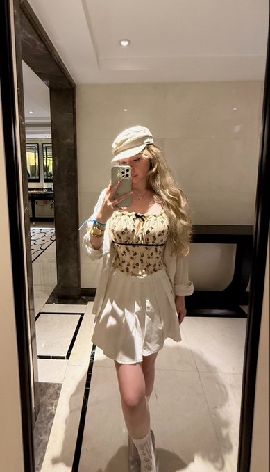 A mirror selfie of a woman wearing a white newsboy cap, a floral corset top over a white dress, and white boots, holding her phone to take the photo in what appears to be a hotel or upscale hallway.