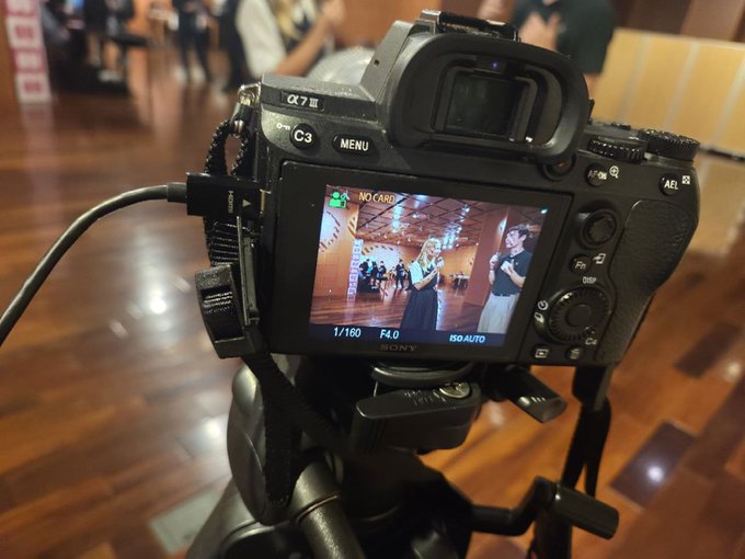 A Sony α7 III camera on a tripod displaying a live view on its rear screen, capturing a woman in a blue dress performing in what appears to be an event venue.