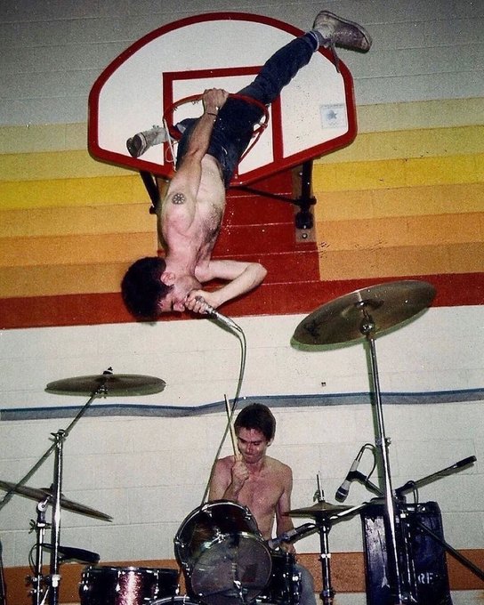 Two shirtless young men appear in a gymnasium, with one hanging upside down from a basketball hoop while the other sits at a drum set below.
