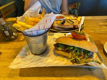 A meal featuring a burger with melted cheese, fresh greens, and tomato is served in a small metal bucket alongside crispy fries and a grilled meat dish on a wooden table. The caption "Lunch date – Closest I'm getting to Carne Asada today" reflects the diner's casual lunch experience, suggesting the burger and grilled meat are their substitute for the traditional Mexican dish.
