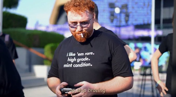A man with red hair and glasses wearing a black t-shirt that reads 'i like 'em rare, high grade, mint condition.', holding a controller with the subtitle 'to stop bracing' below.