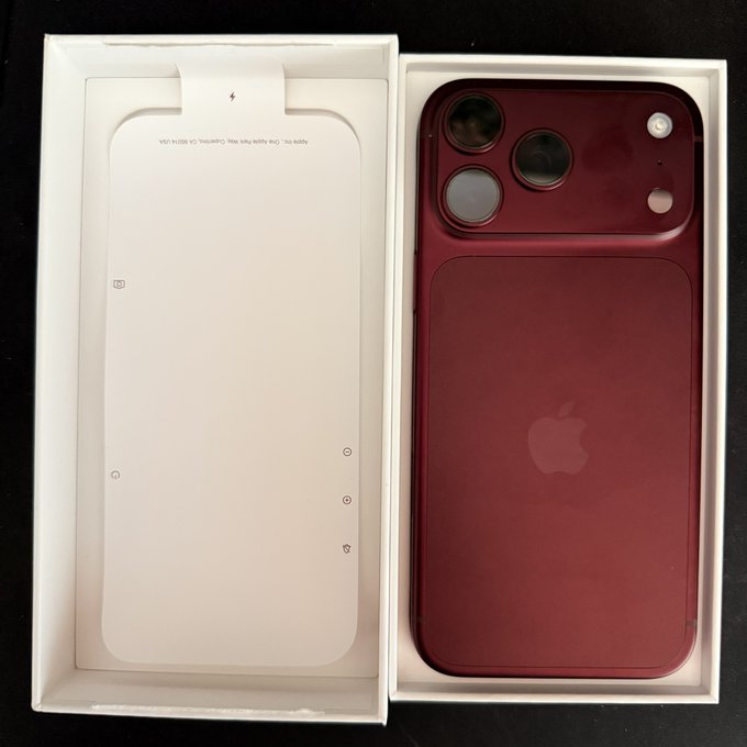 An unboxed new Apple iPhone in a dark red/burgundy color displayed in its white packaging, showing the rear camera module with three lenses and the Apple logo, photographed on a dark surface.