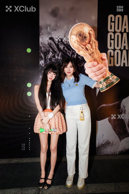 Two young women pose together in front of an XClub backdrop displaying a golden FIFA World Cup trophy and the text 'GOA GOA GOA', with one wearing a plaid skirt and the other in a blue polo shirt.