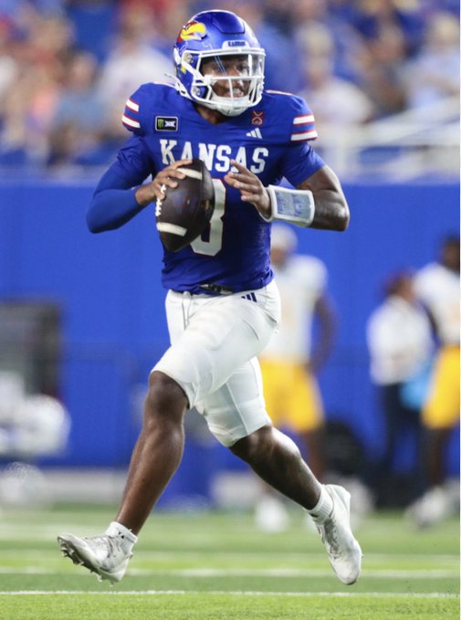 Kansas Jayhawks quarterback #5, Jalon Daniels, in a blue "KANSAS" jersey, drops back to pass while scanning the field during a game. The image aligns with the post's celebration of @KUAthletics, showcasing Jayhawks football in action, with $XRP likely representing the team's "Rock Chalk" crypto-themed community spirit.