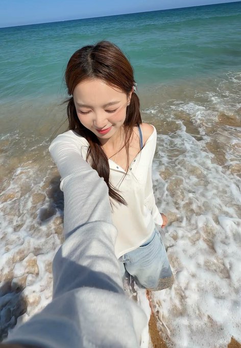 A young woman with long brown hair wearing a white long-sleeve top and denim shorts takes a selfie while standing in the shallow surf of a turquoise sea.