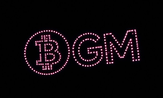 An illuminated pink LED sign on a black background displays the Bitcoin symbol followed by the letters 'GMA'.