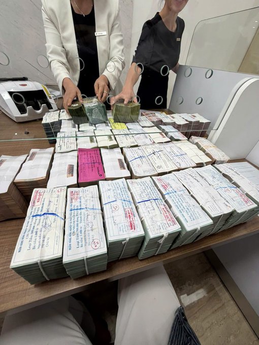 Two bank employees are counting and bundling large stacks of Vietnamese currency (including bundles labeled "NGÂN HÀNG NHÀ NƯỚC VIỆT NAM" - State Bank of Vietnam) at a teller counter, illustrating the post's musing about choosing the "safe" path of savings deposits over other business ventures.