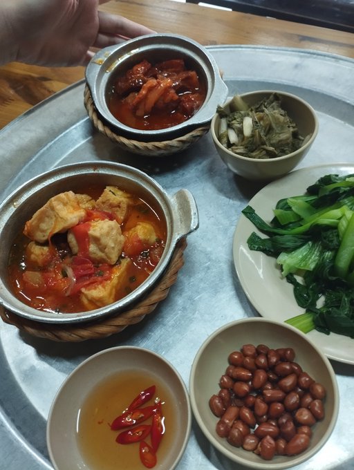 A metal tray holding a meal with tofu stew in tomato sauce, stir-fried greens, peanuts, and chili sauce.