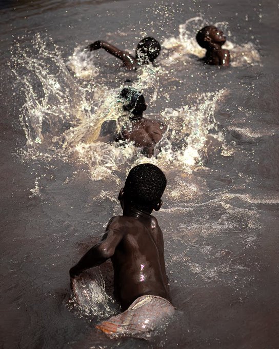 A photograph of three young children playing and splashing in muddy water, with one child in the foreground partially submerged and two others visible in the background creating large splashes.