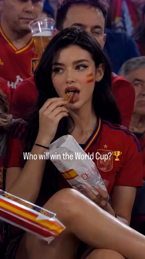 The image features a woman sitting down, eating a snack while watching a soccer game. She has a bag of chips in her hand as she enjoys the match. The background shows other people seated around her, all focused on the game. A text overlay at the bottom of the photo reads "Who will win the World Cup?" and another text that says "6.17梭哈晨报".