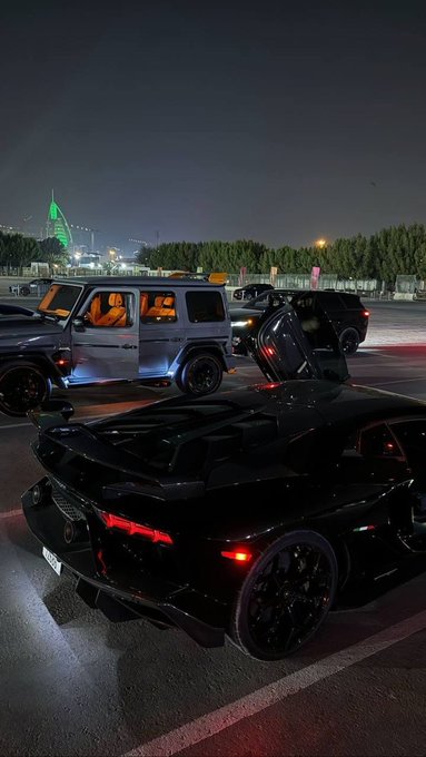 A nighttime street scene shows a sleek black sports car with its scissor door open, parked near a silver Mercedes G-Wagon and other vehicles.