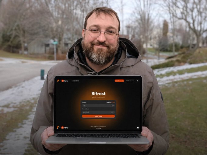 The image shows a man holding up an open laptop computer with the word "Bifrost" displayed on its screen. The man appears to be smiling, possibly indicating that he has found something interesting or useful on the BIFROST platform. The background of the image features trees and a street, suggesting that the man is in an outdoor setting.