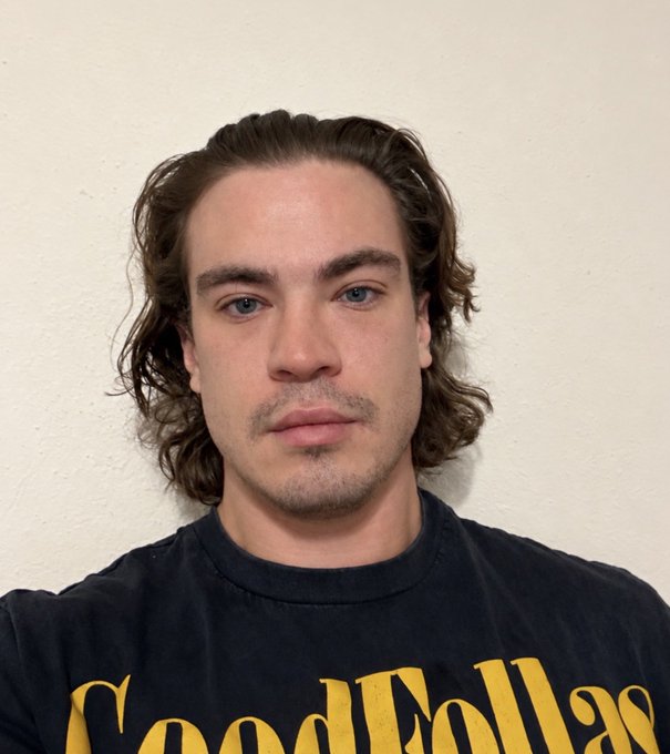 A young man with shoulder-length wavy brown hair and a neutral expression looks directly at the camera, wearing a dark t-shirt with "Good Fellas" printed in yellow text. The image appears to be a selfie taken against a plain white wall, matching the post's plea to be let into another country by presenting a candid, hopeful look.