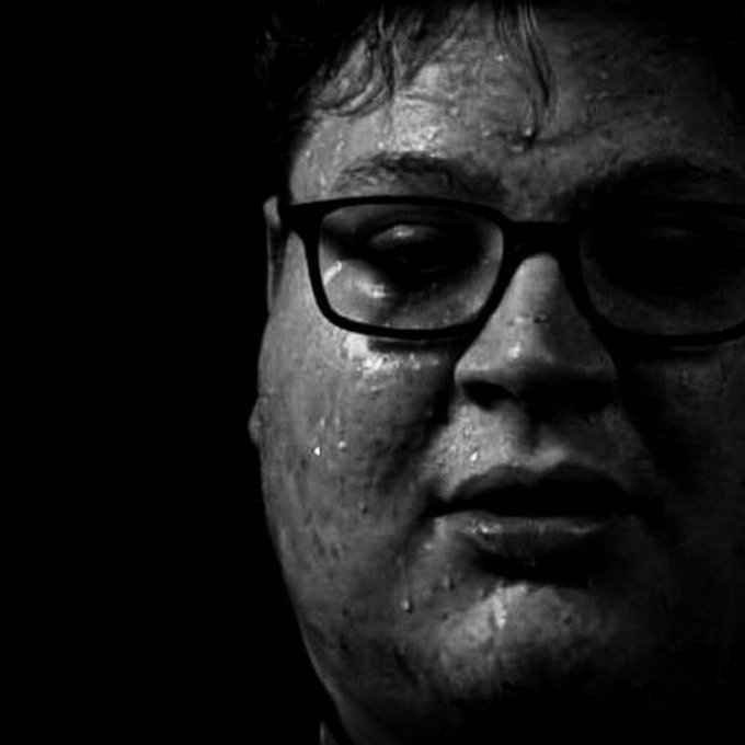 Black and white close-up portrait of a man wearing glasses, his face covered in sweat, set against a dark background.